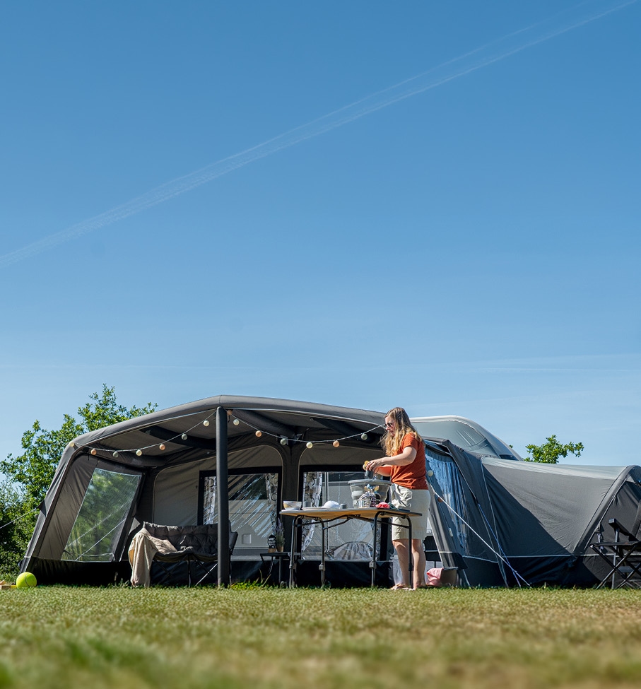 Español: Un amplio avance gris para camper con toldo integrado está montado sobre un césped. Bajo el toldo hay una mesa donde una mujer cocina al aire libre. El avance está decorado con iluminación y asientos, con un cielo azul despejado de fondo.